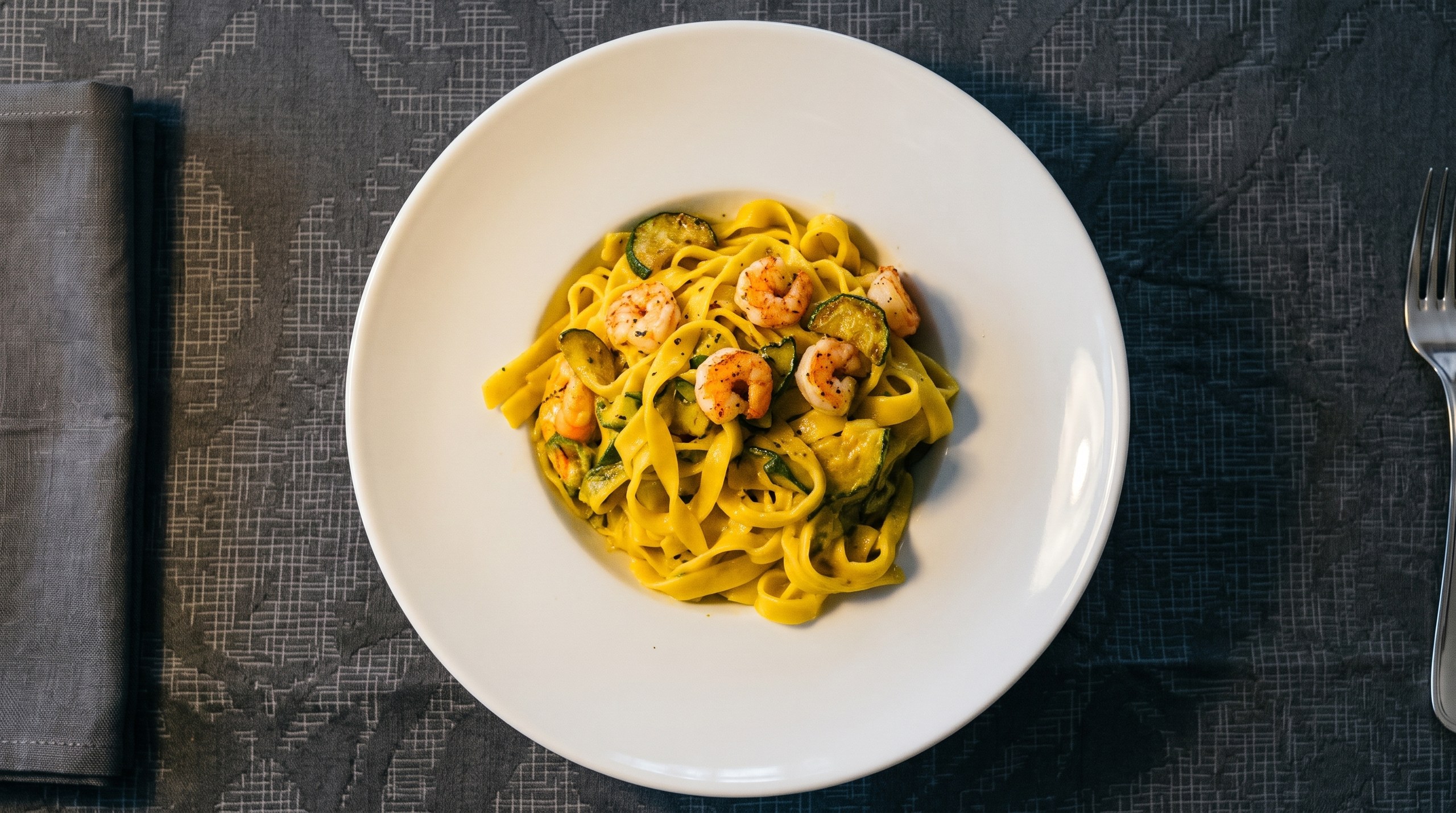 Piatto di tagliatelle allo zafferano con gamberi e zucchine, visto dall'alto su tovaglia grigia